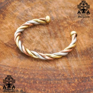 Könnte beinhalten: Ein gedrehtes Metallarmband mit drei verschiedenen Farbbändern aus Silber, Gold und Kupfer. Das Armband ist offen und hat abgerundete Enden.