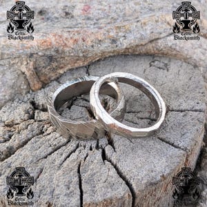 May include: Two silver wedding bands with a hammered finish, resting on a piece of weathered wood. The bands are slightly overlapping, showcasing their unique texture.