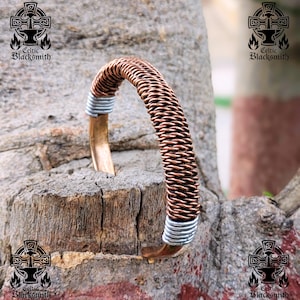 Może przedstawiać: Bransoletka w kolorze miedzi o plecionym wzorze. Bransoletka ma srebrne akcenty na każdym końcu. Obraz zawiera również tekst "Celtic Blacksmith" i logo z krzyżem i kowadłem.