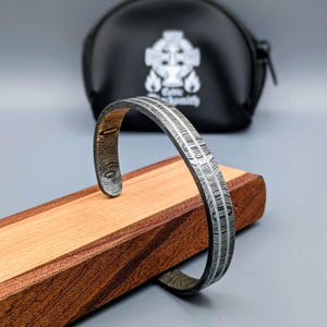 May include: A silver-coloured cuff bracelet with a textured, linear pattern, displayed on a wooden stand. The bracelet has an open design. A black leather pouch with a Celtic cross logo is in the background. Jewellery.