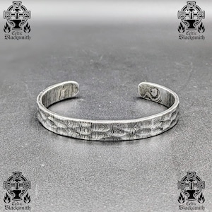 Puede incluir: Un brazalete plateado con una superficie texturizada. El brazalete abierto es de metal. El logotipo de Celtic Blacksmith es visible en las esquinas de la imagen.