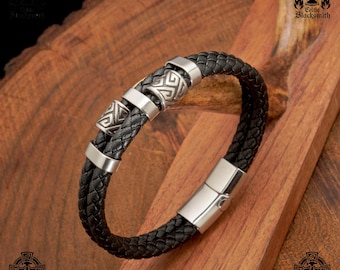 May include: A black leather bracelet with a silver clasp and three silver beads with Celtic knot designs. The bracelet is on a wooden surface.