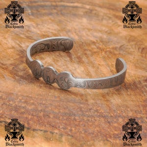 May include: A silver-toned cuff bracelet with a unique wave pattern design. The bracelet has a curved shape and is open at the ends. The surface has a textured, layered appearance. The Celtic Blacksmith logo is visible in the corners.