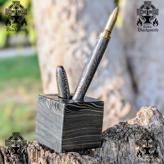 A hand-forged Damascus steel cube with swirling watered-steel patterns doubles as a pen holder and paperweight — 55 favorites don't lie, this thing is stunning.