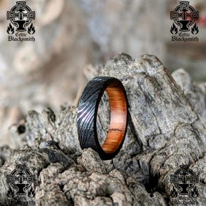 May include: A handcrafted ring with a dark, patterned exterior and a warm, wooden interior. The ring is set against a textured, grey background. The ring is from Celtic Blacksmith.
