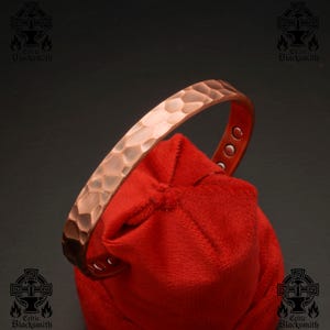 Pode incluir: Uma pulseira de cobre com textura martelada e fechos magnéticos. A pulseira está sobre uma bolsa de veludo vermelho.