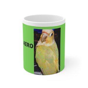 Könnte beinhalten: Eine weiße Keramiktasse mit grünem Hintergrund und einem Bild eines gelben und grünen Papageis mit dem Wort "AERO" auf der Tasse.