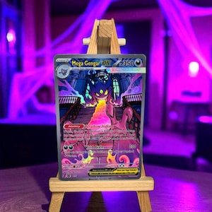 Op de afbeelding: Een Mega Gengar EX Pokémon-kaart wordt getoond op een kleine houten ezel. De kaart toont een paarse en zwarte illustratie van Mega Gengar, met de tekst "Mega Gengar EX" en "350" zichtbaar. De achtergrond is een wazig paars.