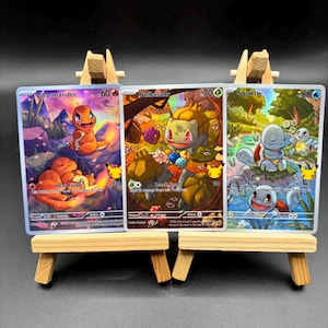 Può includere: Tre carte collezionabili Pokémon esposte su piccoli cavalletti di legno. Le carte presentano Charmander, Bulbasaur e Squirtle, ognuno con opere d'arte e informazioni di gioco uniche. Le carte sono su uno sfondo scuro.