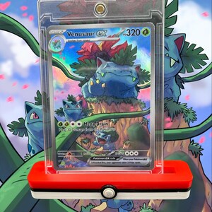 Peut inclure: Une carte Pokémon Venusaur EX dans un étui de protection transparent, présentée sur un support rouge et blanc. La carte présente une illustration de Venusaur bleu avec le texte "Mega Drain" et "320". L'arrière-plan a un design de dessin animé avec d'autres Pokémon.