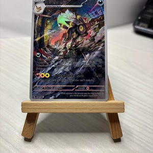 Może przedstawiać: Karta kolekcjonerska Pokémon Umbreon, wyświetlana na małym drewnianym sztaludze. Karta przedstawia czarno-żółtą postać Umbreona z tekstem "Umbreon" i "110". Karta ma opisy ataków i kolorowe tło.