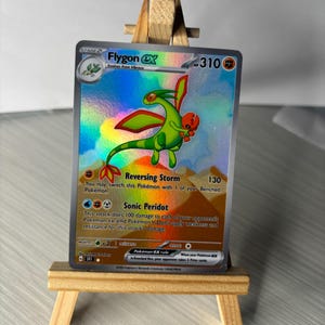 Puede incluir: Una tarjeta Pokémon Flygon ex holográfica, exhibida en un pequeño caballete de madera. La tarjeta presenta una criatura verde y roja con el texto "Flygon ex" y "Stage 2". La tarjeta tiene 310 HP e incluye descripciones de ataque.