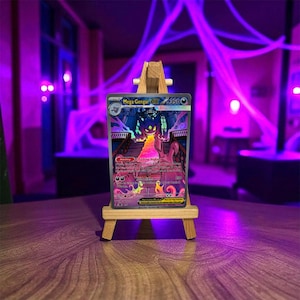 Könnte beinhalten: Eine Mega Gengar Pokémon-Sammelkarte, präsentiert auf einer kleinen Holzstaffelei. Die Karte zeigt eine farbenfrohe Illustration eines Spukhauses und eines lila Geistes. Der Text auf der Karte lautet "Mega Gengar" und "3500".