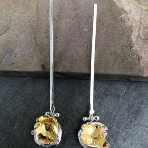 May include: A pair of long, silver earrings with gold accents and a textured, irregular shape at the bottom. The earrings are hanging from a smooth, silver rod.