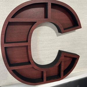 May include: A letter C shaped wooden display shelf with a dark reddish-brown finish and black interior edges. The structure features multiple rectangular compartments separated by thin dividers, creating a grid-like storage space within the letter.