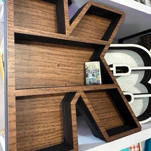 May include: A large wooden letter K shelf with a natural brown wood grain finish and black interior edges. The letter-shaped display unit features multiple horizontal shelves and contains a small book titled The Witness by Nora Roberts.