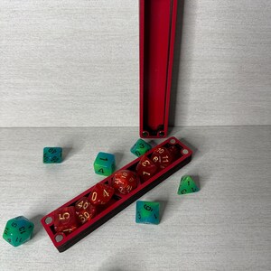 May include: A long, rectangular, black and red wooden dice box with magnetic closures, shown open with one section standing upright. Several translucent red and green polyhedral dice with gold numbering are scattered around the box.