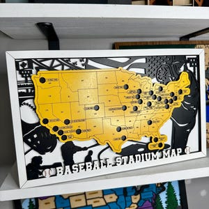 May include: A rectangular framed wall art piece featuring a yellow map of the United States with black circular cutouts for baseball stadiums. The text BASEBALL STADIUM MAP is printed at the bottom, flanked by two small white baseball icons.