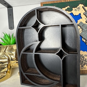 May include: A dark brown wooden display shelf with an arched top and a layered, cut-out design featuring a crescent moon shape and four-pointed star accents. The shelf has multiple internal compartments created by the layered wood structure.