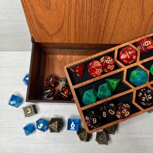 May include: A wooden box with a removable tray containing various multi-sided polyhedral dice in red, green, blue, and black colors with gold numbering. Additional loose dice in blue, black, and red are scattered around the base of the box.
