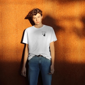 Könnte beinhalten: Ein weißes T-Shirt mit einem schwarzen grafischen Aufdruck einer Person, die eine Einkaufstasche hält. Der Text auf dem Aufdruck lautet "It's a Tee Thing".