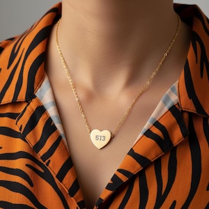 May include: A gold-colored necklace with a heart-shaped pendant. The pendant has the number "513" engraved on it. The necklace is worn against a background of an orange and black patterned shirt.