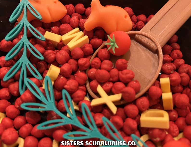 Puede incluir: Un contenedor sensorial lleno de cuentas rojas, formas de peces y zanahorias naranjas, bloques amarillos con forma de queso, un tomate rojo y una cuchara beige. Formas de &aacute;rboles y hojas turquesas est&aacute;n dispersas. El texto "SISTERS SCHOOLHOUSE CO." est&aacute; en la parte inferior.