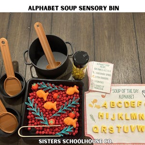 Puede incluir: Caja sensorial de sopa de letras con una variedad de alimentos y herramientas de juego. Incluye una olla negra, sartenes peque&ntilde;as y cucharas naranjas. Una bandeja contiene cuentas rojas, galletas con forma de pez y tomates. El alfabeto se muestra con pasta en forma de letras.