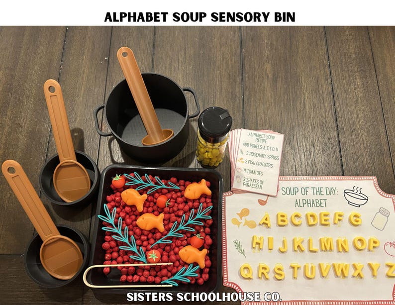 Puede incluir: Caja sensorial de sopa de letras con una variedad de alimentos y herramientas de juego. Incluye una olla negra, sartenes peque&ntilde;as y cucharas naranjas. Una bandeja contiene cuentas rojas, galletas con forma de pez y tomates. El alfabeto se muestra con pasta en forma de letras.