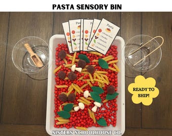 May include: A sensory bin labeled "PASTA SENSORY BIN" filled with red beads, penne pasta, felt leaves, and felt meatballs. Includes small bowls, a wooden scoop, and tongs. The bin has cards with a pasta recipe. A yellow sign says "READY TO SHIP!"