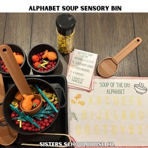 Puede incluir: Un contenedor sensorial con tem&aacute;tica de sopa de letras. Incluye una olla negra, cuencos y cucharas llenas de alimentos de juguete coloridos como tomates, galletas de pescado y pasta de letras. Tambi&eacute;n se incluye una tarjeta de recetas y un letrero con el alfabeto.