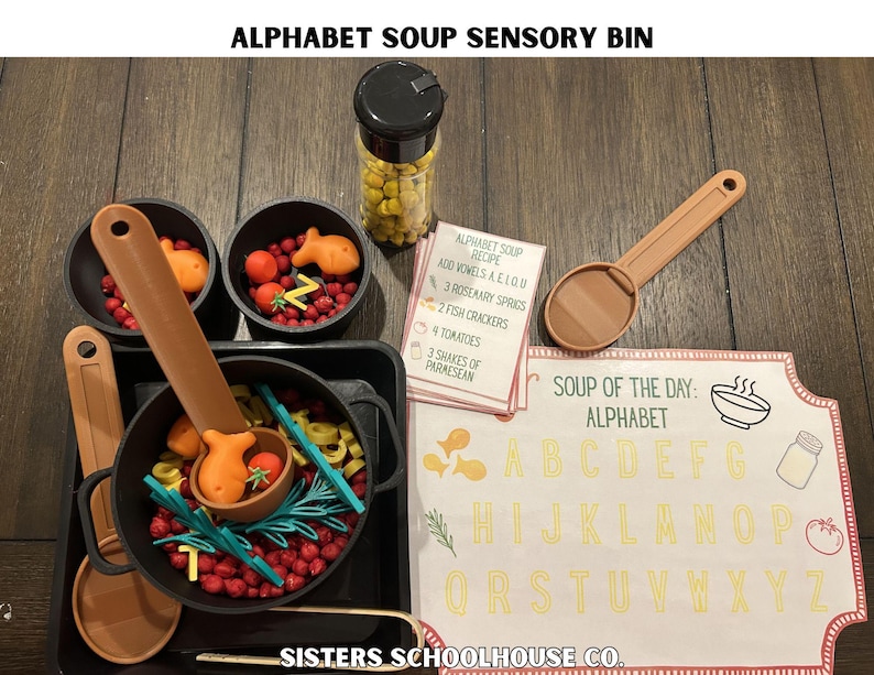 Puede incluir: Un contenedor sensorial con tem&aacute;tica de sopa de letras. Incluye una olla negra, cuencos y cucharas llenas de alimentos de juguete coloridos como tomates, galletas de pescado y pasta de letras. Tambi&eacute;n se incluye una tarjeta de recetas y un letrero con el alfabeto.