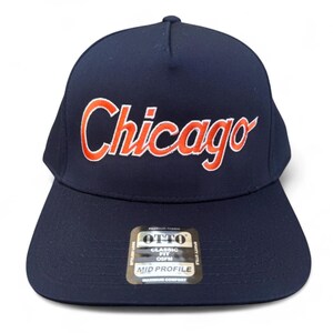 Może przedstawiać: Granatowa czapka z daszkiem z napisem "Chicago" wyhaftowanym pomarańczową czcionką. Czapka ma klasyczny krój i średni profil. Dołączona jest etykieta z informacjami o produkcie.