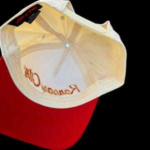 May include: A baseball cap with a red peak and a cream-coloured crown. The interior is white with gold embroidered text that reads "Kansas City". The cap has a snapback closure.