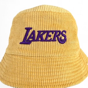 Puede incluir: Sombrero de pescador de pana amarillo mostaza con la palabra "LAKERS" bordada en morado. El sombrero tiene una corona redondeada y un ala inclinada hacia abajo. La tela de pana tiene estrías verticales.