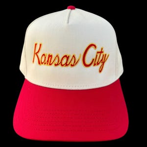 May include: A baseball cap in white and red, featuring the text "Kansas City" embroidered in gold script. The cap has a red peak and a red button on top. A classic style hat.