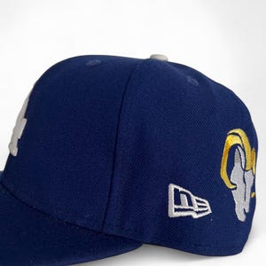May include: A navy blue baseball cap with a curved brim. The cap features a white embroidered logo on the front and a gold and gray ram logo on the side. The New Era logo is embroidered in white on the side.