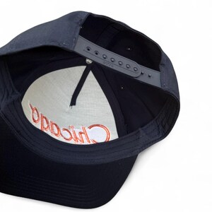 May include: Navy blue baseball cap with a gray adjustable strap. The interior features a white panel with the word "Chicago" embroidered in red. The cap is viewed from the underside, showcasing the stitching and construction details.