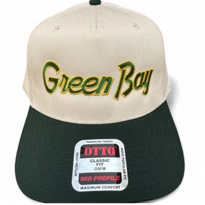 May include: A beige and dark green baseball cap with the text "Green Bay" embroidered in green. The cap has a flat brim and a tag that reads "OTTO CLASSIC FIT OSFM MID PROFILE MAXIMUM COMFORT".