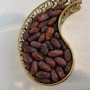 May include: A gold-coloured, crescent-shaped serving dish filled with numerous brown dates. The dish has a decorative, openwork design along its rim, featuring silhouettes of mosques and stars. The dates are oval-shaped and have a wrinkled texture.