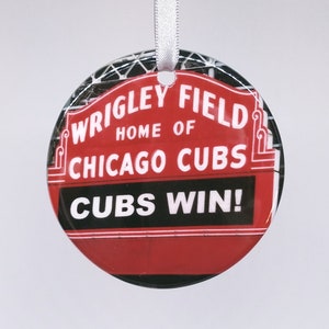 May include: Round ornament featuring the Wrigley Field sign, home of the Chicago Cubs. The ornament is red with white text that reads "Wrigley Field Home of Chicago Cubs" and "Cubs Win!" A white ribbon is attached for hanging.
