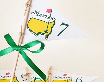 Puede incluir: Un conjunto de 5 banderas de golf verdes y blancas con un contorno amarillo de los Estados Unidos y la palabra "Masters" impresa en cada bandera. Las banderas están numeradas del 5 al 9. Cada bandera está unida a un palo de madera con una cinta verde atada alrededor.