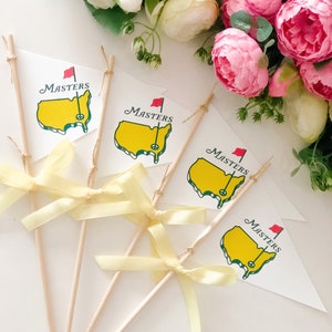 Könnte beinhalten: Vier weiße Flaggen mit gelben und grünen Golfplatzdesigns und dem Wort "Masters" auf jeder Flagge. Die Flaggen sind an Holzstöcken mit gelben Schleifen befestigt.