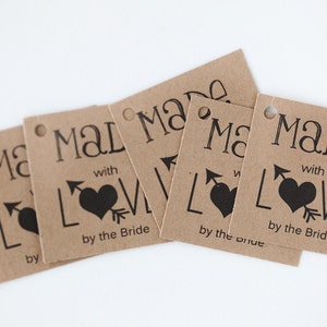 Puede incluir: Cinco etiquetas de regalo de papel marr&oacute;n con el texto "Made with Love by the Bride" y un coraz&oacute;n negro con una flecha que lo atraviesa.