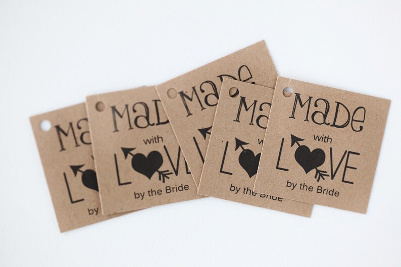 Puede incluir: Cinco etiquetas de regalo de papel marr&oacute;n con el texto "Made with Love by the Bride" y un coraz&oacute;n negro con una flecha que lo atraviesa.