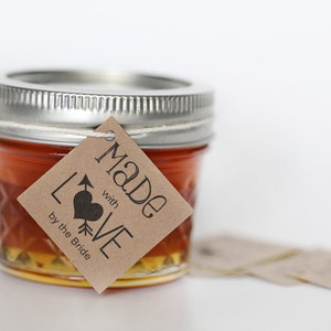 Puede incluir: Un peque&ntilde;o frasco de vidrio lleno de un l&iacute;quido dorado, con una etiqueta de papel marr&oacute;n atada alrededor de la tapa. La etiqueta dice "Made with LOVE by the Bride".