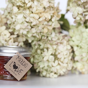 Puede incluir: Un peque&ntilde;o frasco de vidrio lleno de un l&iacute;quido rojo, con una etiqueta de papel marr&oacute;n que dice "Made with LOVE by the Bride". El frasco est&aacute; sentado frente a un ramo de hortensias blancas.