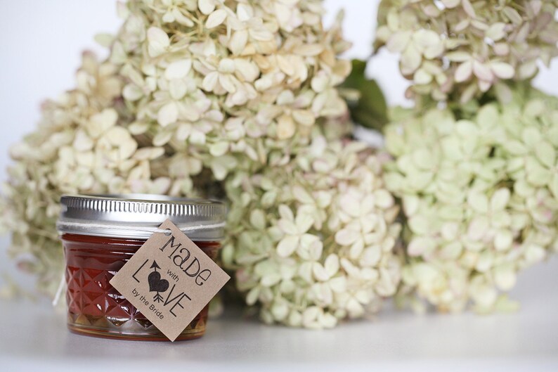 Puede incluir: Un peque&ntilde;o frasco de vidrio lleno de un l&iacute;quido rojo, con una etiqueta de papel marr&oacute;n que dice "Made with LOVE by the Bride". El frasco est&aacute; sentado frente a un ramo de hortensias blancas.