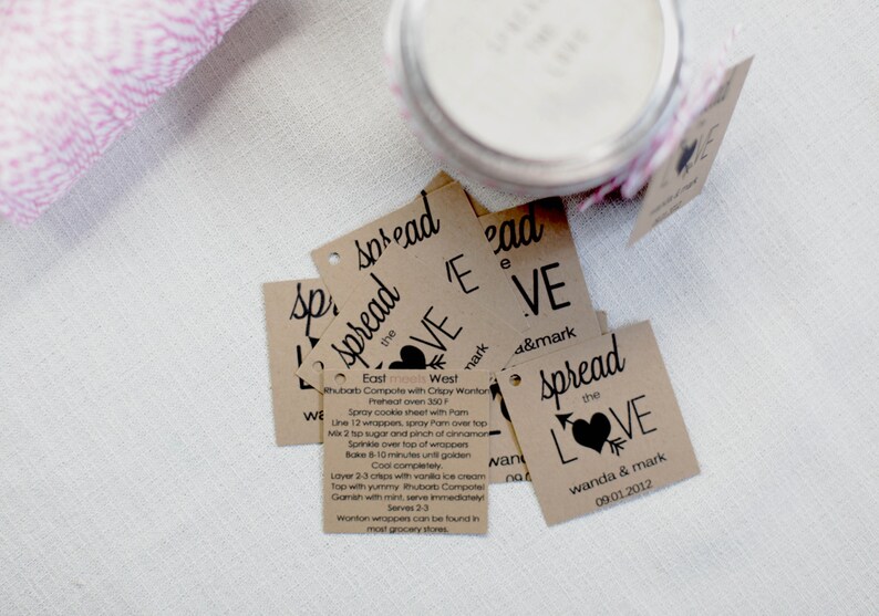 Puede incluir: Un conjunto de etiquetas de regalo de papel marr&oacute;n con texto negro y un dise&ntilde;o de coraz&oacute;n. Las etiquetas dicen "Spread the Love" y "Wanda & Mark 09.01.2012".