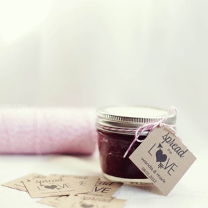 Puede incluir: Un peque&ntilde;o tarro de vidrio lleno de mermelada roja oscura, atado con un cordel y una etiqueta de papel marr&oacute;n que dice "Spread the Love" con un coraz&oacute;n y una flecha. La etiqueta tambi&eacute;n incluye los nombres "Wanda & Mark" y la fecha "09.01.2012".